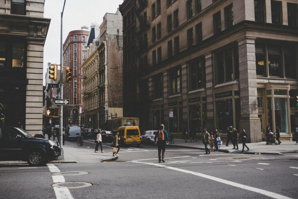 A bustling street scene in Manhattan, featuring pedestrians crossing the street, a yellow taxi, and classic New York City buildings lining the sidewalks.