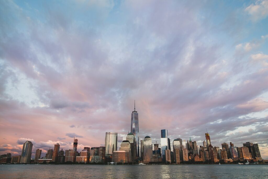 A panoramic view of the New York City skyline at sunset, featuring the iconic One World Trade Center and surrounding skyscrapers, with the Hudson River in the foreground. The image captures the essence of the city's luxurious and expensive neighborhoods, highlighting the architectural grandeur and urban landscape.