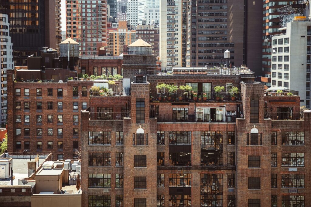 A multi-story apartment building with a modern design, featuring large windows and rooftop gardens, set against an urban backdrop with other high-rise buildings visible in the distance.