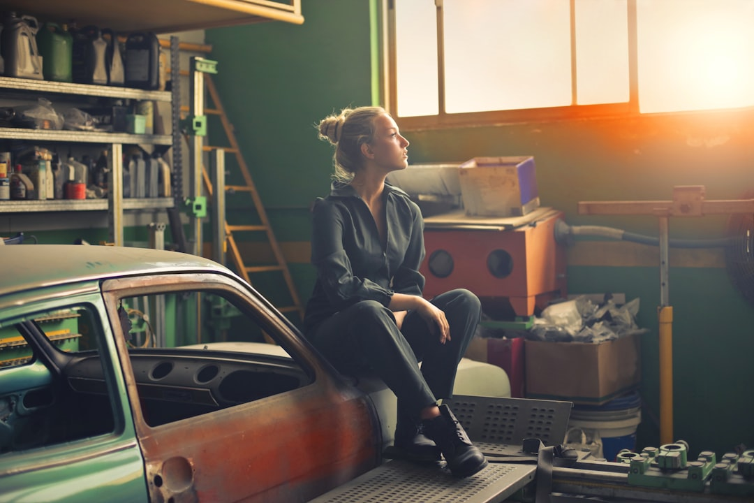 A woman sitting on the edge of a vintage car in a well-lit garage, surrounded by shelves filled with tools and equipment, looking thoughtfully into the distance.