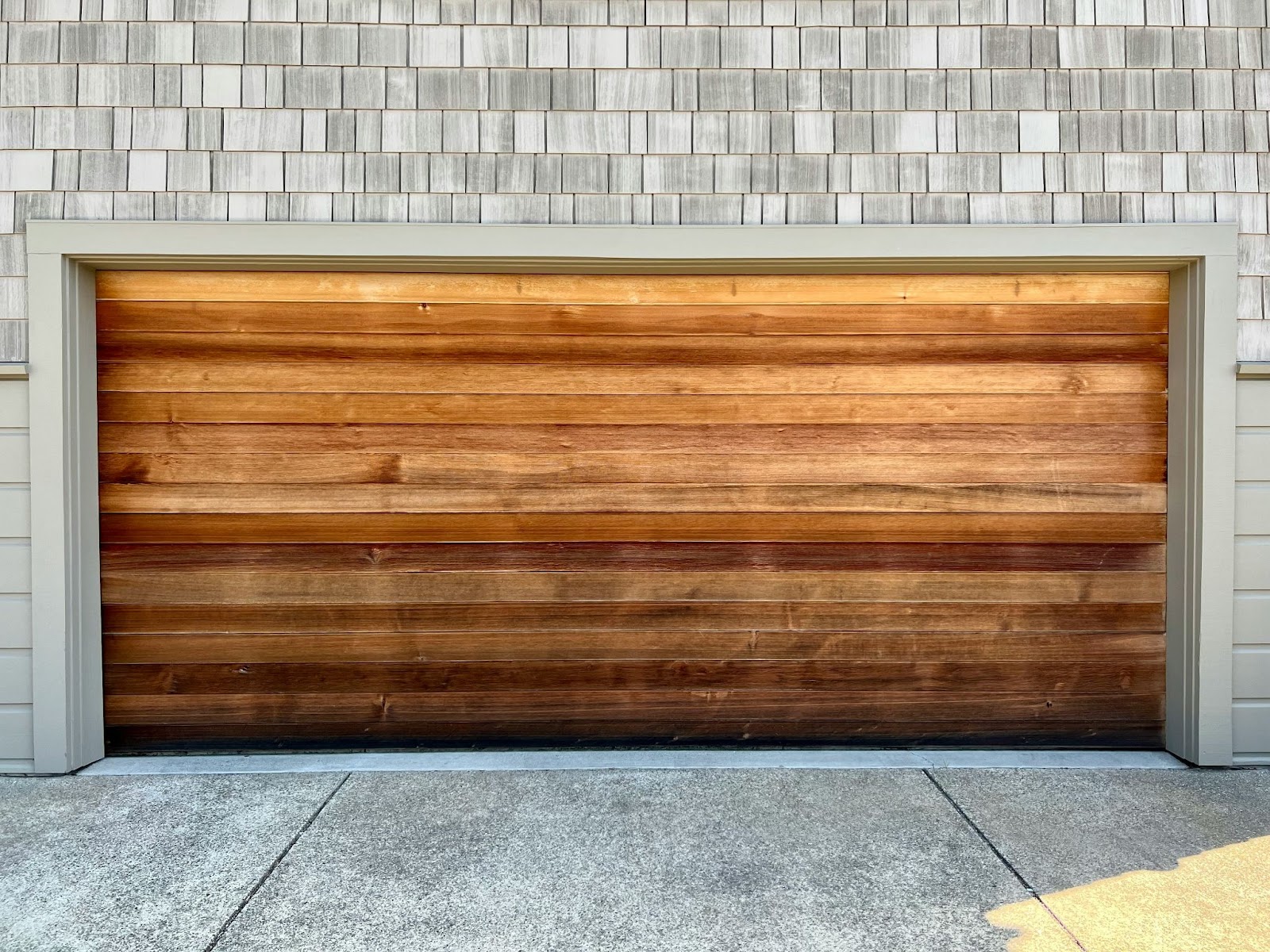 A closed wooden garage door with horizontal panels, set in a light-colored brick wall with a shingled upper section, and a concrete driveway in front.