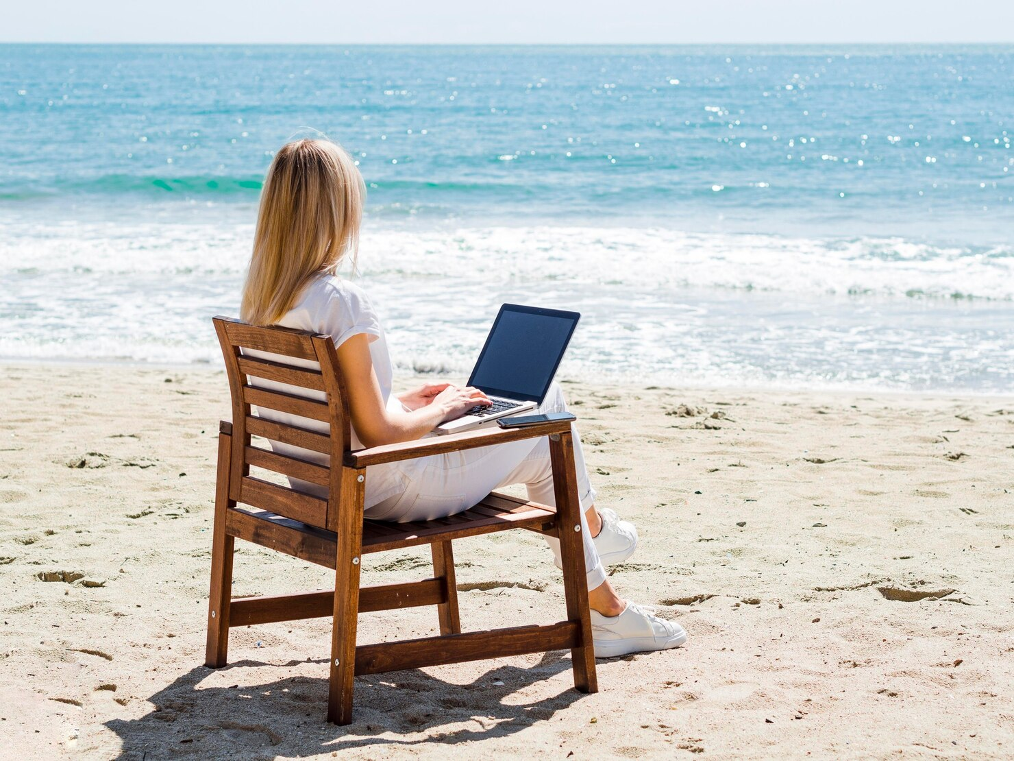 A woman with a laptop sitting on a chair at a beach 