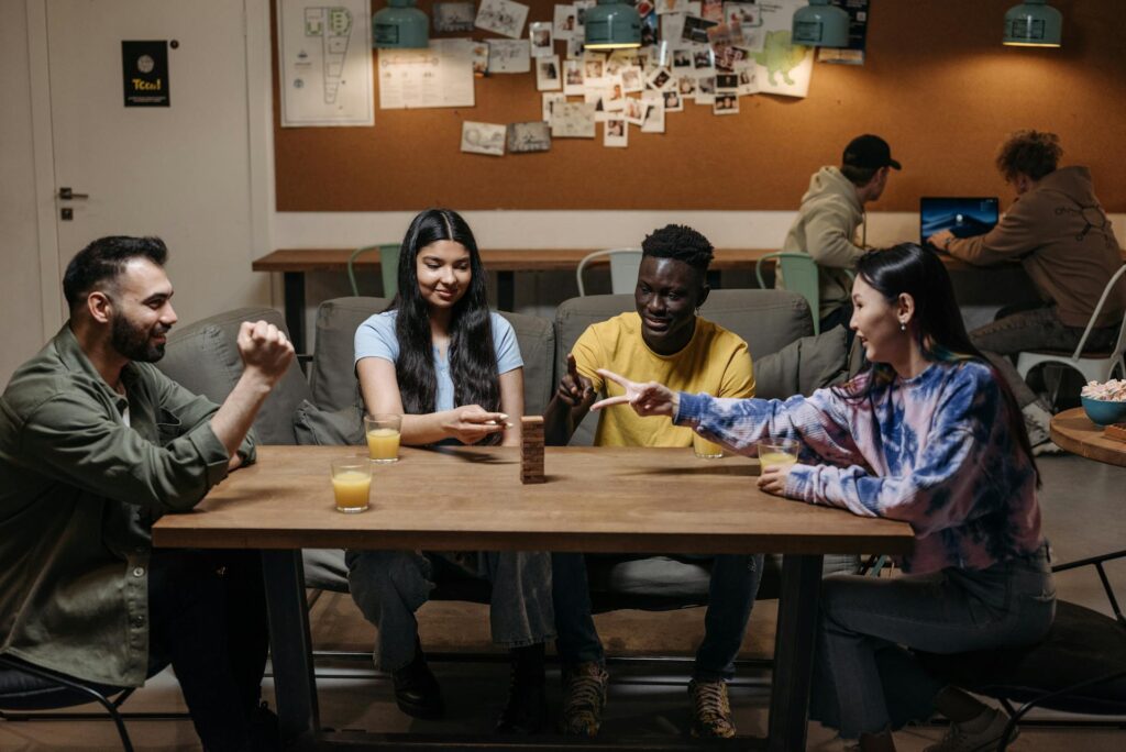 Four friends sit around a table playing Jenga in a cozy café or lounge, smiling and interacting while drinks sit on the table, with other people working in the background.